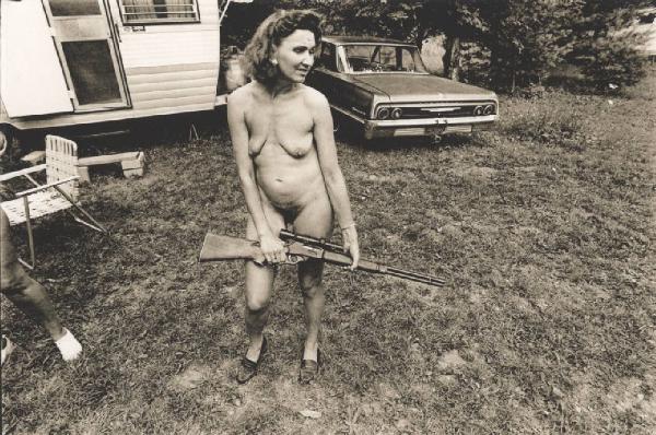 Donna nuda con fucile posa in giardino davanti a una roulotte