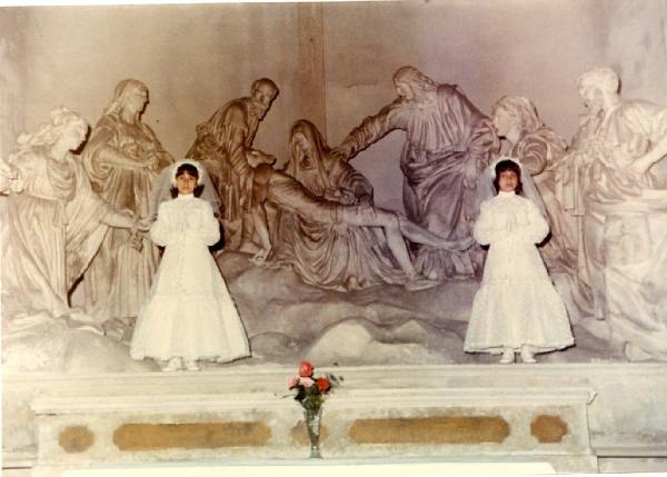Due ragazzine posano con vestito della prima comunione. Altare. Vaso di fiori. Sculture religiose. Rappresentazione della deposizione di Cristo