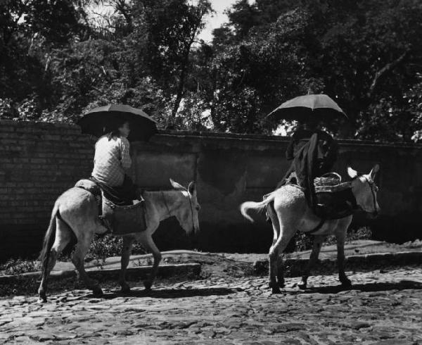 Coppia di donne contadine sopra due asini nei pressi di un muro di cinta