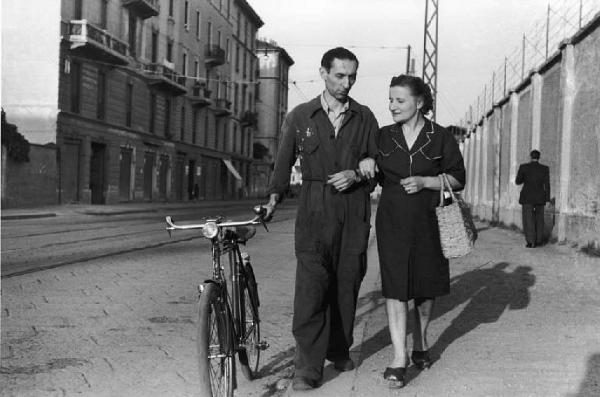 Italia Dopoguerra. Milano - Strada - Ritratto di coppia: donna a braccetto con un uomo in tuta da operaio - Bicicletta