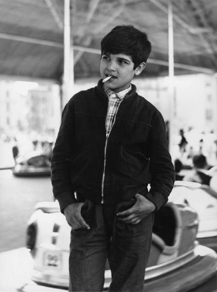Luna park. Milano - Luna park - Ritratto infantile - Bambino con sigaretta in bocca sul bordo della pista degli autoscontri - Giostra sullo sfondo - Fumo
