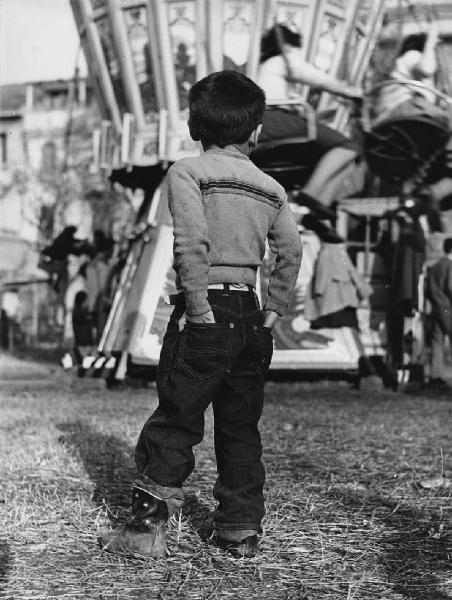 Luna park. Milano - Luna park - Ritratto infantile - Bambino di spalle con le mani in tasca - Jeans e stivali - Giostre sullo sfondo