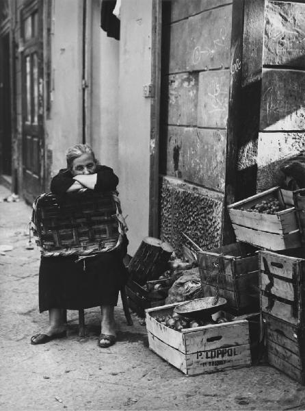 Napoli: Riposo. Napoli - Vicoli - Ritratto femminile - Venditore ambulante di frutta e ortaggi: donna anziana su una sedia con cesta in mano - Riposo