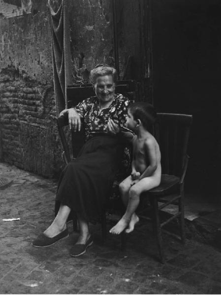 Napoli: Nei Bassi. Napoli - Vicoli - Anziana con bambino nudo seduti su delle sedie davanti all'uscio di casa