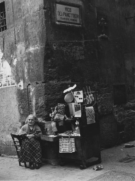 Napoli: Si vende si compra. Napoli - Vicoli - Vico dei Panettieri Quartiere Pendino - Ritratto femminile - Anziana seduta su una sedia: venditore ambulante - Banchetto con merci, oggetti