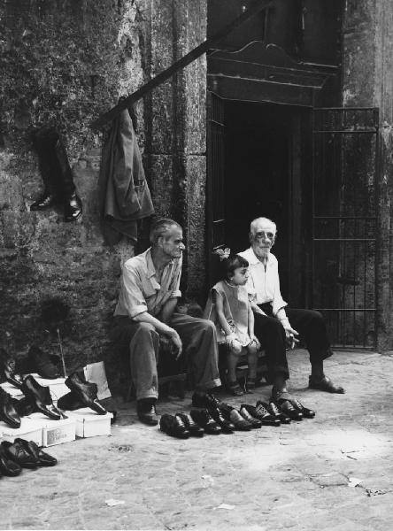Napoli: Si vende si compra. Napoli - Vicoli - Ritratto di gruppo - Anziani con bambina seduti su una panca, venditori ambulanti di scarpe