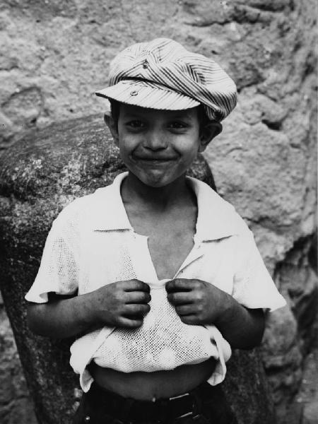 Napoli (?): Volti figure, ragazzi. Napoli (?) - Esterno - Ritratto infantile - Bambino con cappello e maglietta alzata piena di oggetti - Sorriso