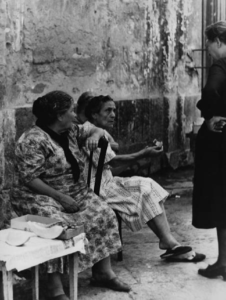 Napoli: Conversazioni. Napoli - Vicoli - Anziane sedute su sedie braccio teso verso anziana in piedi