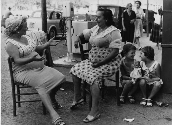 Napoli: Conversazioni. Napoli - Pompa di benzina - Ritratto di gruppo - Anziane sedute su sedie con braccia aperte e coppia di bambine con bambole sedute su un gradino