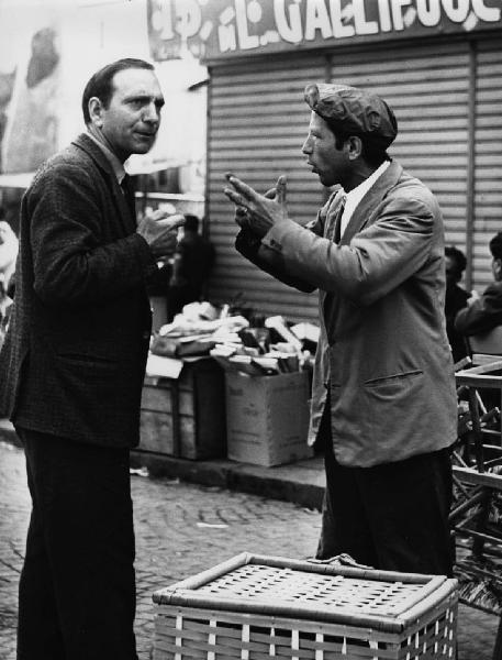 Napoli: Conversazioni. Napoli - Vicoli - Ritratto maschile - Uomo venditore ambulante parla con altro uomo - Gestualità delle mani