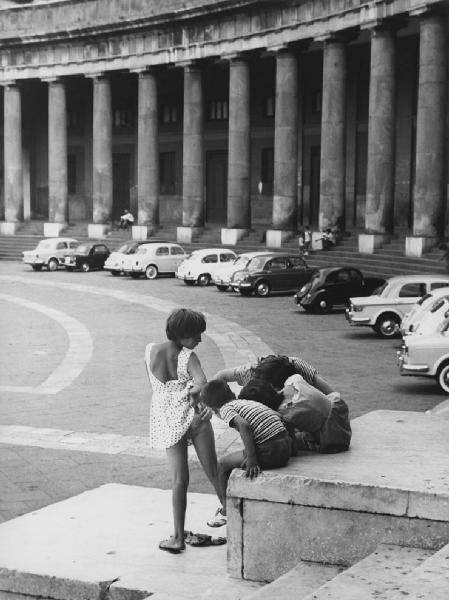 Napoli: Scene di vita varie. Napoli - Piazza del Plebiscito - Ritratto di gruppo - Bambina con vestito alzato verso due bambini e adulto - Automobili FIAT sullo sfondo