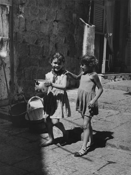 Napoli: Scene di vita varie. Napoli - Vicoli - Ritratto infantile - Bambina con pentola in mano e un'altra le poggia la mano sulla spalla - Sorrisi