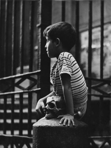 Napoli: Scene di vita varie. Napoli - Vicoli - Ritratto infantile - Bambino seduto su un paracarro con teschio in bronzo