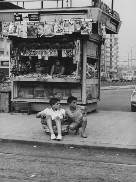 Napoli: Scene di vita varie. Napoli - Strada, marciapiede - Ritratto infantile - Bambino seduto in una cesta di vimini con a fianco un altro bambino - Edicola in secondo piano