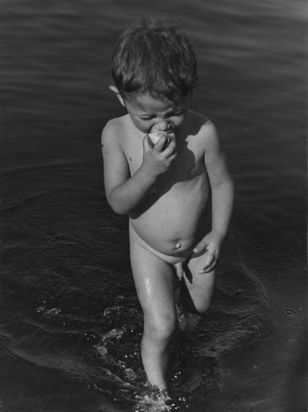 Napoli: Bimbi, soli. Napoli - Mare - Ritratto infantile - Bambino nudo nell'acqua mangia un frutto