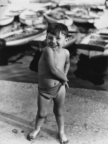 Napoli: Bimbi, soli. Napoli - Mare - Banchina - Ritratto infantile - Bambino in costume da bagno - Sorriso - Barche sfocate sullo sfondo