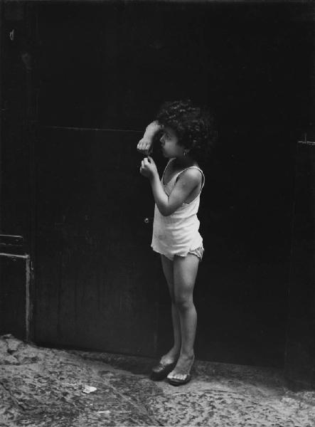 Napoli: Bimbi, soli. Napoli - Strada - Ritratto infantile - Bambina in intimo e canottiera con scarpe da donna sull'uscio di casa