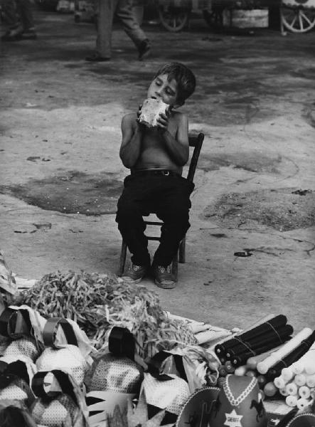 Napoli: Bimbi, soli. Napoli - Strada - Ritratto infantile - Bambino seduto su una sedia con pane in bocca - Bancarella con maschere e oggetti di carnevale