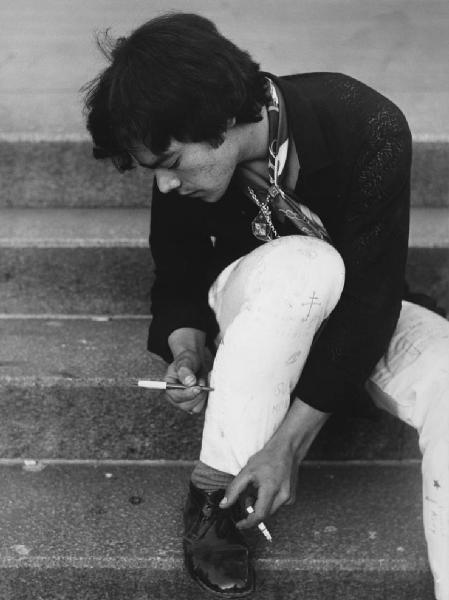 Mondo Beat. Milano - Piazza del Duomo - Ritratto maschile - Ragazzo seduto sui gradini scrive a penna sui pantaloni, jeans