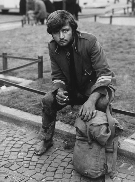 Mondo Beat. Milano - Piazza del Duomo - Ritratto maschile - Ragazzo seduto su ringhiera dell'aiuola: giacca militare, a piedi nudi, sigaretta, zaino