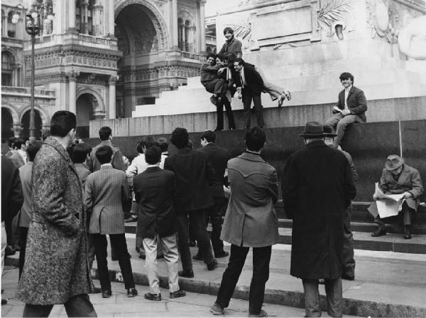 Mondo Beat. Milano - Piazza del Duomo - Monumento a Vittorio Emanuele II - Ragazzi in posa sul monumento - Altri uomini e ragazzi in primo piano di spalle