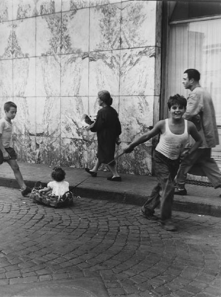 Napoli: Bimbi, scene di vita varie. Napoli - Strada - Bambino tira cesta con bambina - Gioco - Passanti