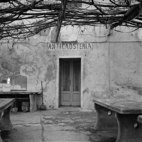 Milano - Peschiera Borromeo - Strada SS415 Paullese - Insegna dipinta con scritta: "Antica Osteria" - Cortile - Copertura con rami - Tavoli in pietra - Porta a vetri con sbarre in ferro