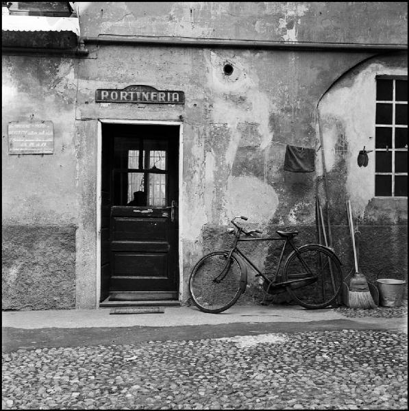 Milano - Via Ascanio Sforza 21 - Portineria, esterno - Bicicletta - Insegna - Attrezzi per la pulizia - Filo per stendere i panni - Silhouette di uomo all'interno della portineria