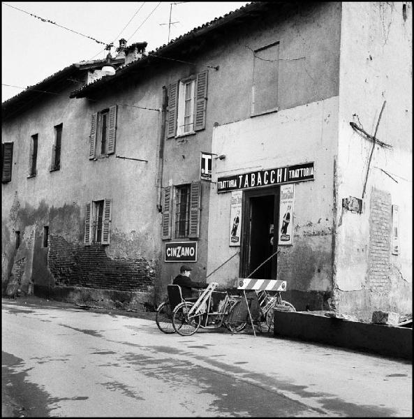 Milano - Via Moncucco 33 - Trattoria/Tabacchi, esterno - Strada - Ritratto maschile: uomo anziano in sedia a rotelle - Biciclette parcheggiate - Insegna Cinzano - Insegne Coca Cola - Croce di rami con ex voto