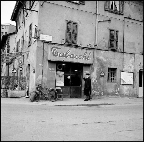 Milano - Via Abbadesse angolo via degli Assereto - Tabacchi, esterno - Ritratto maschile: uomo con bastone da passeggio - Bicicletta parcheggiata - Insegna con scritta: "tabacchi"