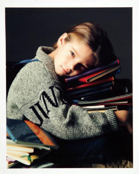 Campagna pubblicitaria per Trussardi Junior - Bambina seduta di profilo con maglione di lana grigio melange - Testa posata su pila di quaderni