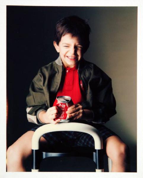 Campagna pubblicitaria per Trussardi Junior - Bambino sorridente seduto: giubbotto verde miltare su polo rossa e bermuda scozzesi - Coca-Cola