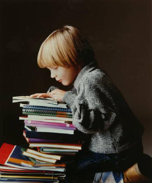 Campagna pubblicitaria per Trussardi Junior - Bambino con pila di quaderni e libri: maglione grigio melange su blue jeans e calze a rombi
