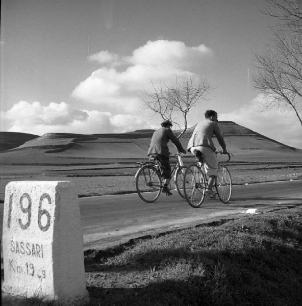 Italia del Sud. Sardegna - strada Macomer-Sassari - pietra miliare con la scritta "196 Sassari km 19.3" - uomini in bicicletta