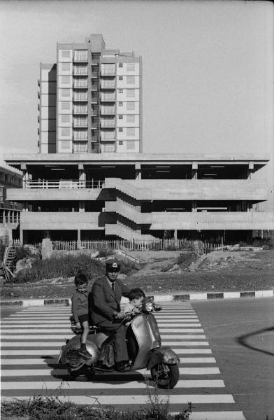 Milano. Periferia - Uomo su uno scooter Vespa con due bambini - Sullo sfondo: edificio in costruzione abbandonato e casa popolare