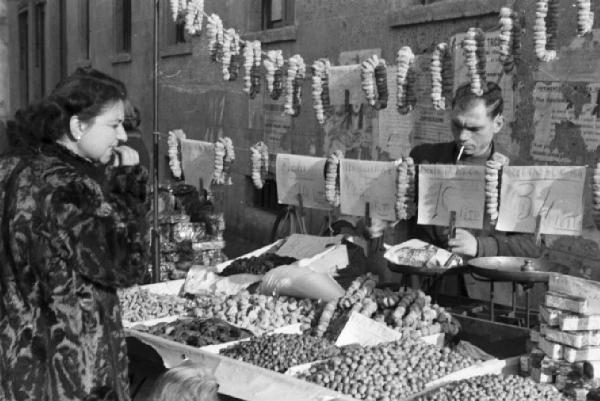 Italia Dopoguerra. Milano - Fiera degli Obèi - Obèi - Banco frutta secca - Donna