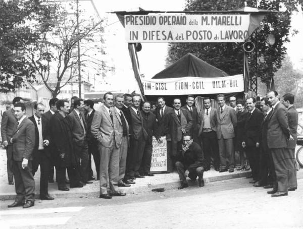 Magneti Marelli - Presidio operaio davanti alla fabbrica in difesa del posto di lavoro - Visita di una delegazione sindacale - Foto di gruppo, tra i sindacalisti L. Banfi, A. Pizzinato, P. Boni, P. Perotta, B.Trentin, A. Breschi - Striscione