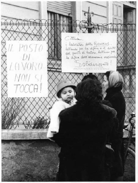 Sciopero dei lavoratori della General di Bresso contro la sospensione a tempo indeterminato - Donna con bambino legge i cartelli di protesta appesi a una recinzione