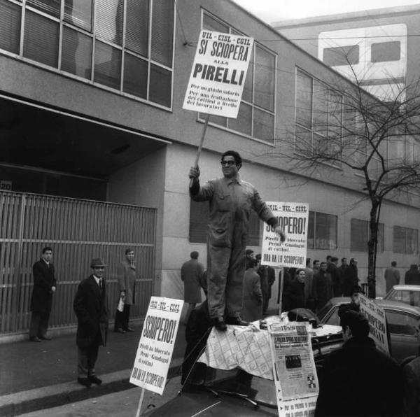 Sciopero dei lavoratori della Pirelli - Operaio sul tetto di un auto mostra cartelli di sciopero davanti alla sede della direzione - Operaio con tuta da lavoro - Cartelli Cgil, Cisl, Uil