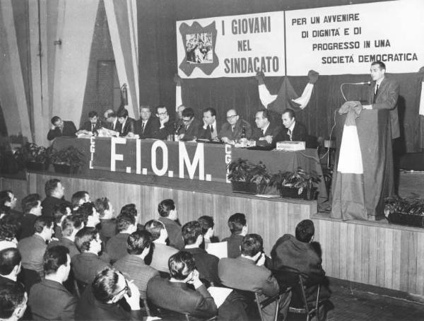 Camera del Lavoro - Salone "Di Vittorio" - Interno - Conferenza provinciale dei giovani metalmeccanici - Palco - Tavolo della presidenza - Antonio Pizzinato al microfono - Parola d'ordine - Striscione Fiom - Bandiera