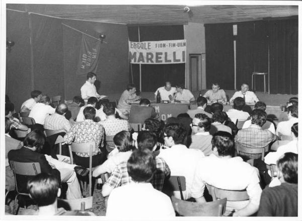 Interno - Assemblea dei lavoratori della Ercole Marelli per il rinnovo del contratto - Panoramica sulla sala - Tavolo della presidenza con Antonio Pizzinato e Bruno Trentin - Platea con pubblico di spalle - Striscione - Bandiera Fim, Fiom, Uilm