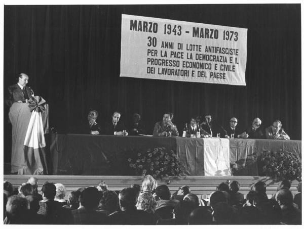 Teatro Odeon - Interno - Commemorazione degli scioperi del marzo 1943 - Antifascismo - Palco - Tavolo della presidenza con tra gli altri R. Romei, M. Colombo, L. De Carlini, P. Perotta - A. Bonaccini al microfono - Parola d'ordine - Bandiera italiana