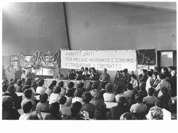 Imperial - Interno - Assemblea permanente dei lavoratori per il contratto - Tavolo della presidenza con tra gli altri A. Japà, P. Boni - Annio Breschi al microfono - Operaie con grembiule da lavoro - Striscione - Parola d'ordine - Bandiera Flm Milano