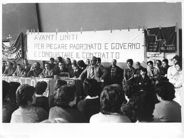 Imperial - Interno - Assemblea permanente dei lavoratori per il contratto - Tavolo della presidenza con tra gli altri A. Breschi e A. Japà - Relatore al microfono - Operaie con grembiule da lavoro - Parola d'ordine - Bandiera Flm Milano