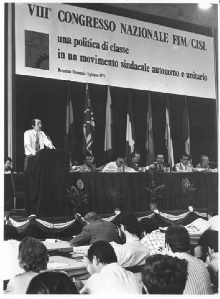 Sala - Interno - VIII congresso nazionale della Fim Cisl - Tavolo della presidenza - Oratore al microfono - Parola d'ordine - Bandiere