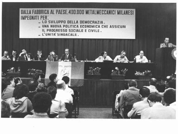 Camera del Lavoro - Salone "Di Vittorio" - Interno - Assemblea Quadri Fiom - Tavolo della presidenza con A. Pizzinato, L. De Carlini, P. Galli, L. Banfi, A. Bellocchio, B. Marabese - Annio Breschi al microfono - Parola d'ordine - Bandiera Fiom