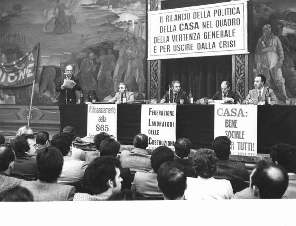 Convegno unitario edili per il problema della casa - Interno - Tavolo della presidenza, tra gli oratori Gustavo Scarpino - Gianfranco Rognoni al microfono - Parola d'ordine - Manifesto - Striscione