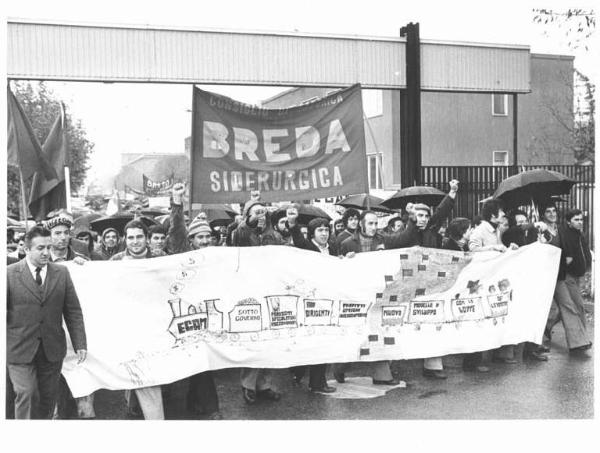 Sciopero dei lavoratori della Breda Siderurgica - Corteo sotto la pioggia esce dalla fabbrica per andare davanti all'Intersind - Operai salutano con il pugno chiuso - Striscioni - Bandiere