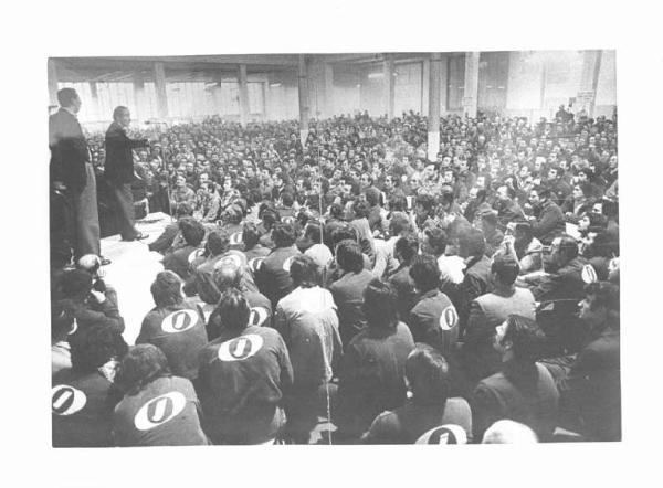 Fabbrica Innocenti Leyland (Innse) - Interno - Assemblea dei lavoratori - Palco - Bruno Storti al microfono, al suo fianco Lucio De Carlini - Platea con operai con tuta da lavoro - Cartelli