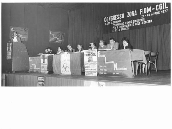 Congresso Fiom Cgil zona Sesto San Giovanni - Interno - Palco - Tavolo della presidenza con, tra gli oratori, Antonio Pizzinato - Giovanni Chinosi al microfono - Parola d'ordine - Bandiera Flm - Manifesti - Bandiera Cgil Camera del Lavoro